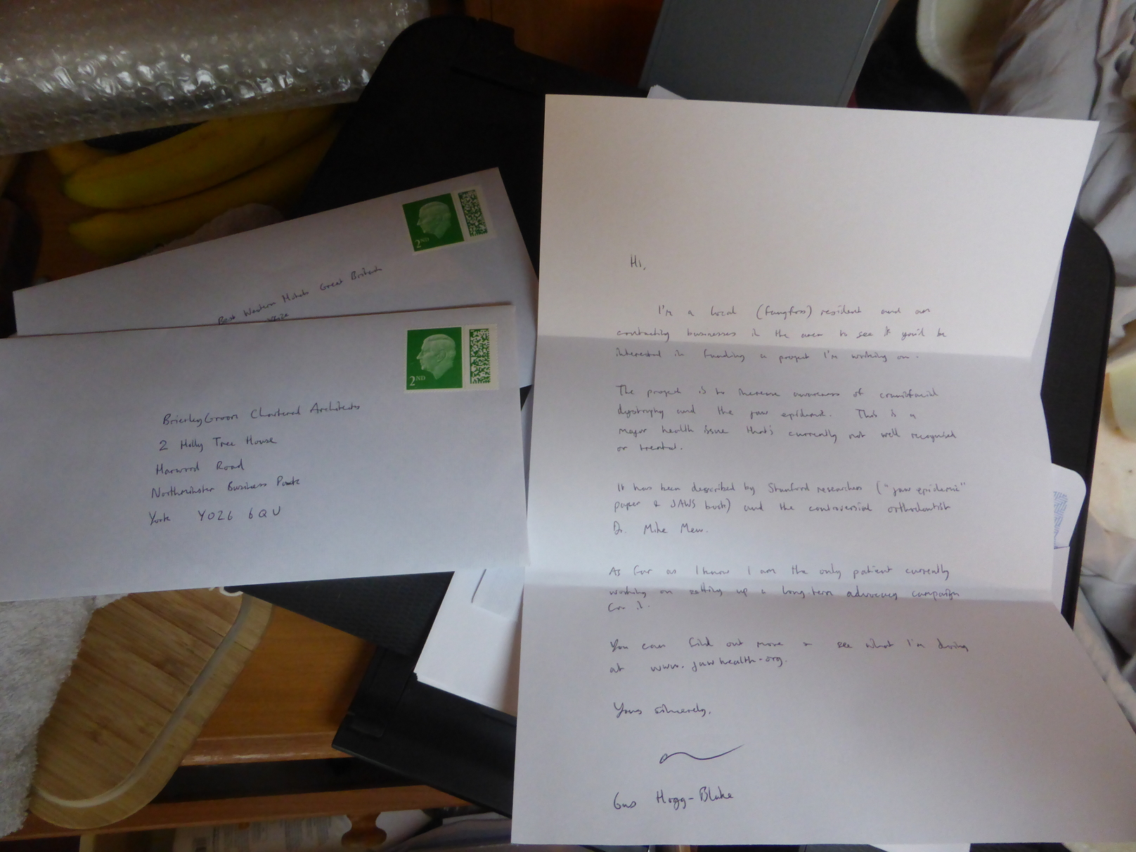 A hand-written letter atop some sealed envelopes with second-class stamps, all atop a printer on a messy surface.